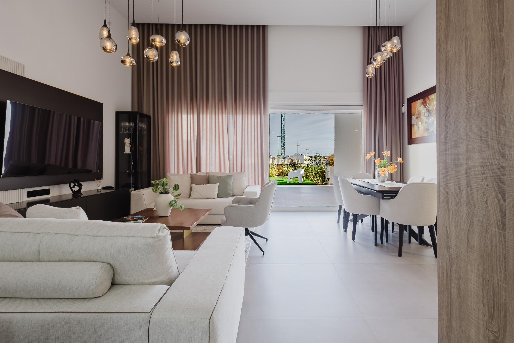 Open-concept living and dining room with cream sofas, pendant lights, and a large window showing a garden outside