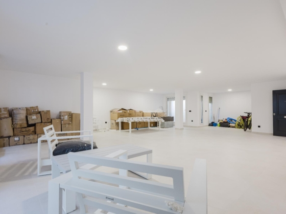 Large, bright unfinished basement/garage with stacked cardboard boxes along left wall and white outdoor furniture in the foreground; dark blue door on the right.