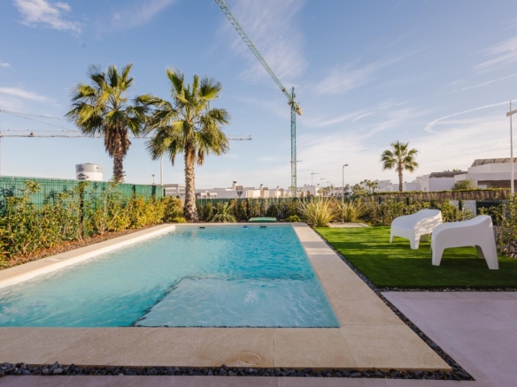 Rectangular swimming pool in a sunny backyard with palm trees, a manicured lawn, and two white chairs; construction cranes visible in the distance behind a green fence.