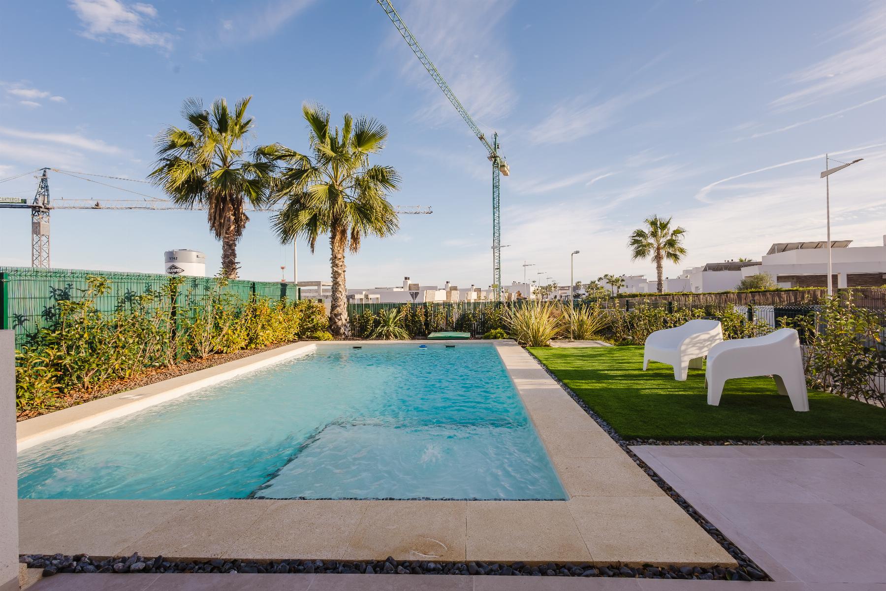 Rectangular swimming pool in a sunny backyard with palm trees, a manicured lawn, and two white chairs; construction cranes visible in the distance behind a green fence.