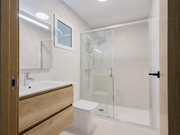 Modern bathroom with a glass shower enclosure, white fixtures, and a light wood vanity against marble walls.