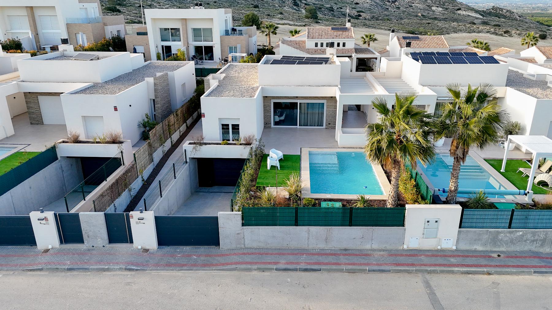 Aerial view of a modern hillside residential complex with white boxy homes and blue swimming pools.0 pensaike