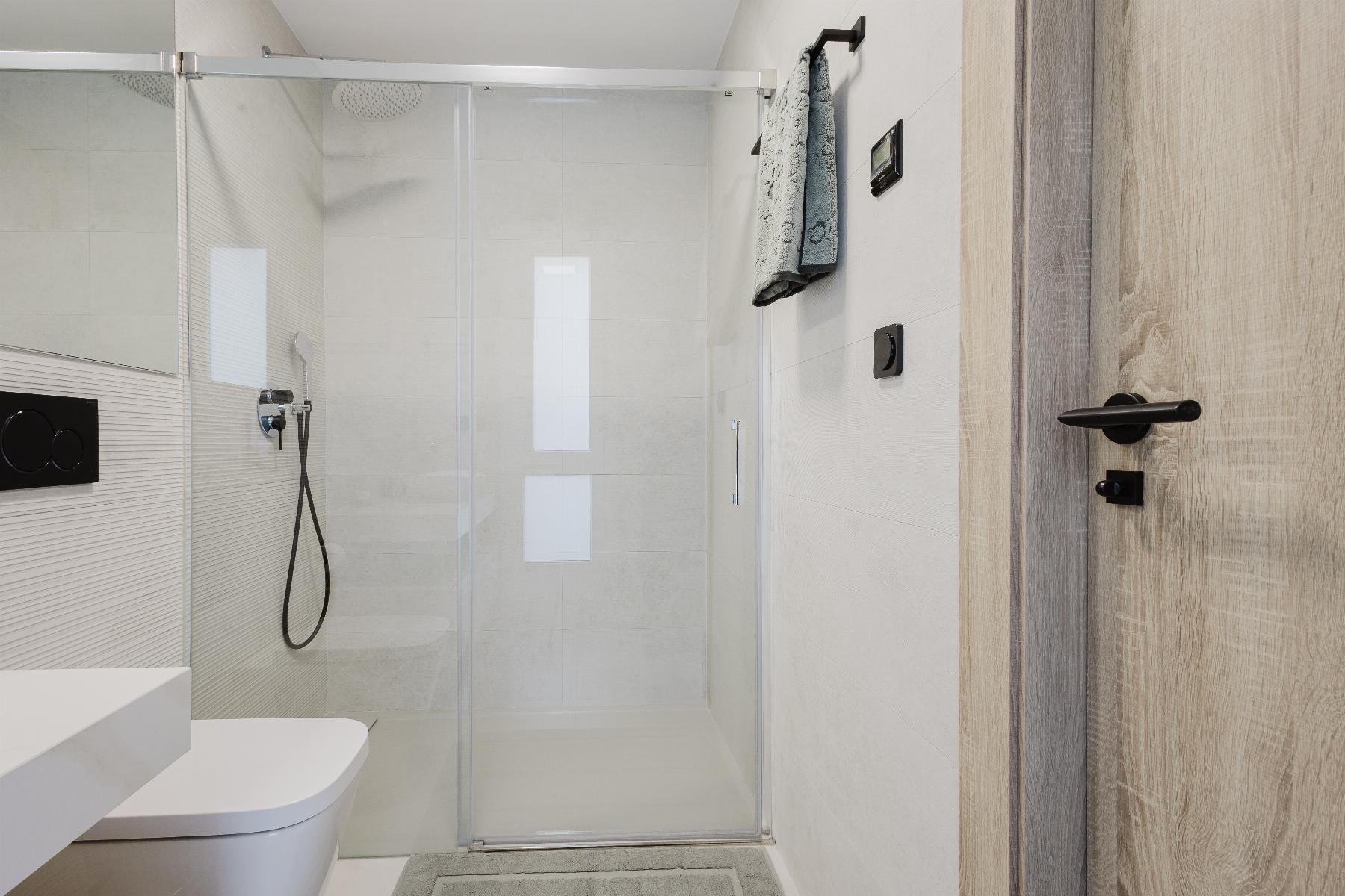 Modern bathroom with a glass shower enclosure, wall-hung toilet, and light wood door on the right.