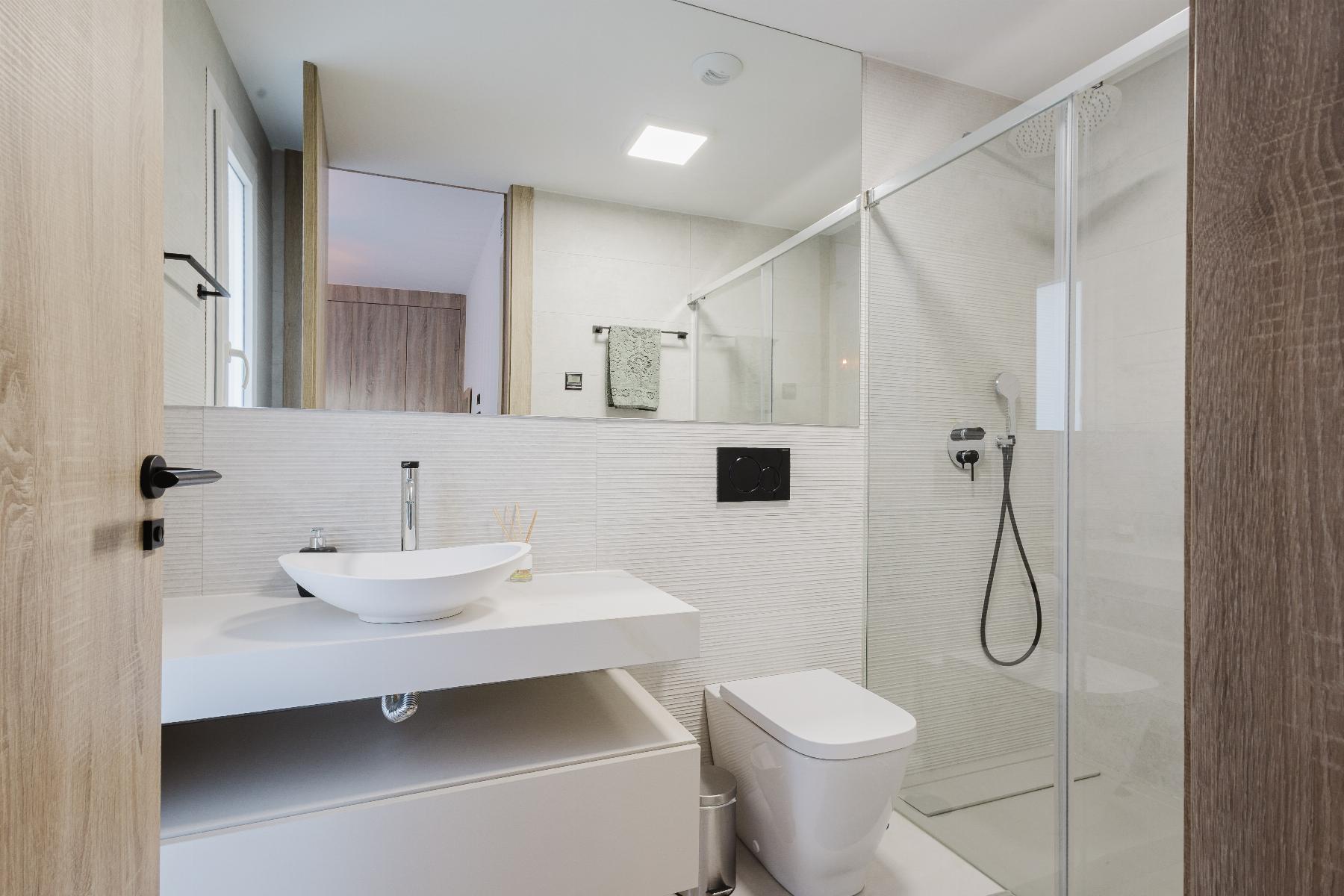 Modern bathroom with wall-mounted vanity, white vessel sink, and chrome faucet facing a large mirror and glass shower enclosure.