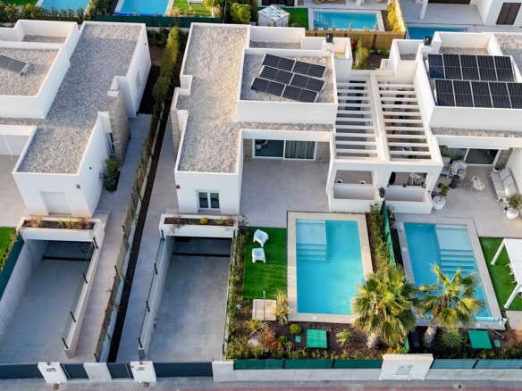 Aerial view of modern white houses with solar panels on flat roofs, blue swimming pools, and green lawns along a gated row of homes