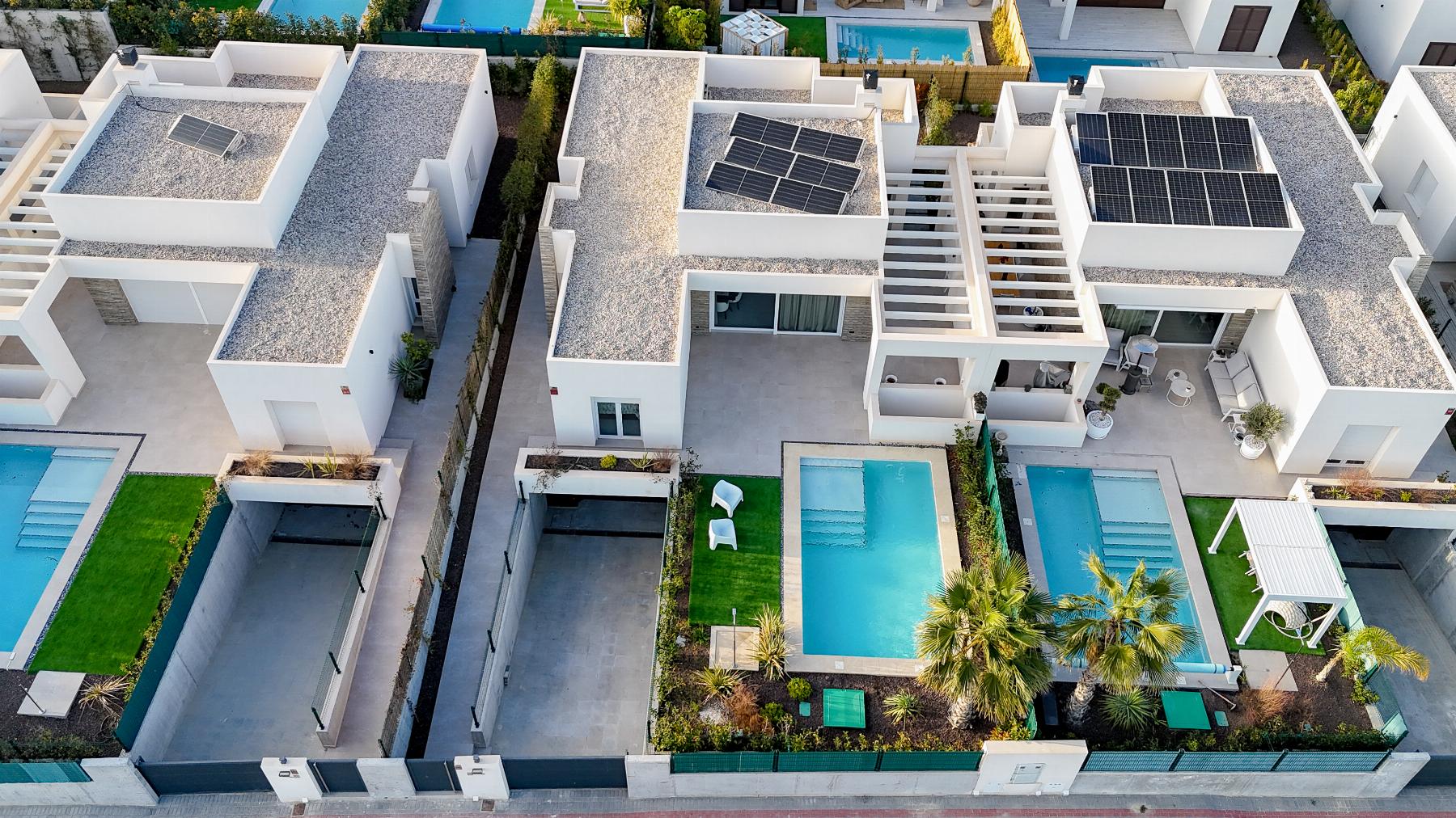 Aerial view of modern white houses with solar panels on flat roofs, blue swimming pools, and green lawns along a gated row of homes