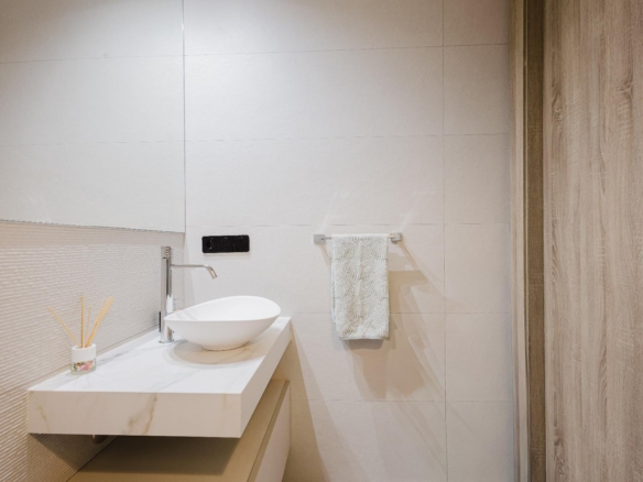 Modern bathroom with a round white vessel sink on a marble countertop, chrome faucet, beige tiled walls, a small reed diffuser, and a wooden door on the right.