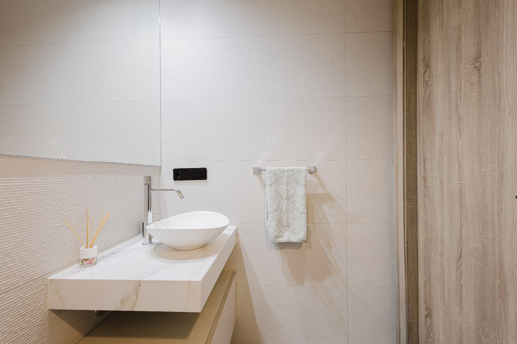 Modern bathroom with a round white vessel sink on a marble countertop, chrome faucet, beige tiled walls, a small reed diffuser, and a wooden door on the right.