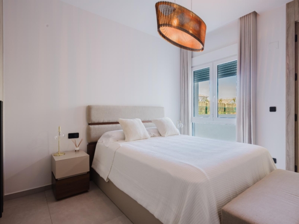 Bright, modern hotel bedroom with a white bed, beige upholstered headboard, nightstand, and warm pendant light; large window with light curtains letting in daylight.