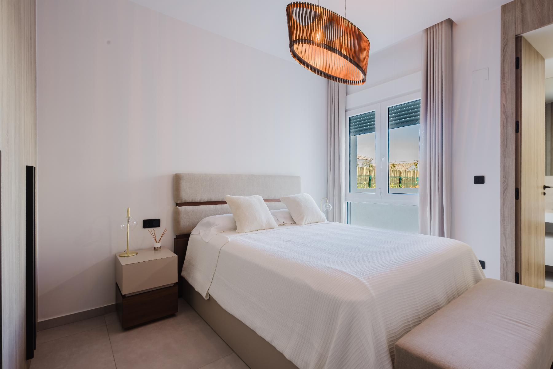 Bright, modern hotel bedroom with a white bed, beige upholstered headboard, nightstand, and warm pendant light; large window with light curtains letting in daylight.