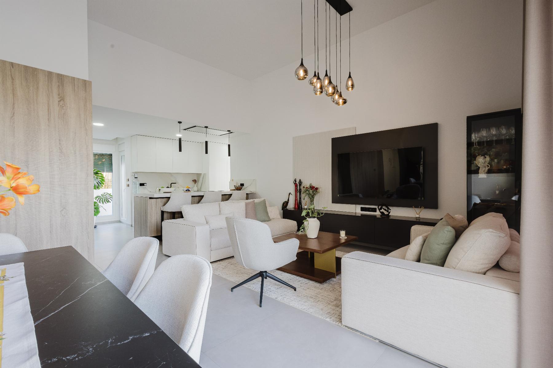 Open-concept living area with beige sofas, a large flat-screen TV on a dark wall panel, and a cluster of hanging pendant lights.