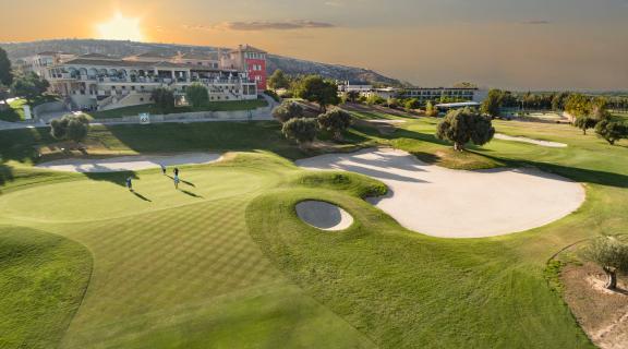 Sunlit golf course with a green, sand bunker, and clubhouse in the background.