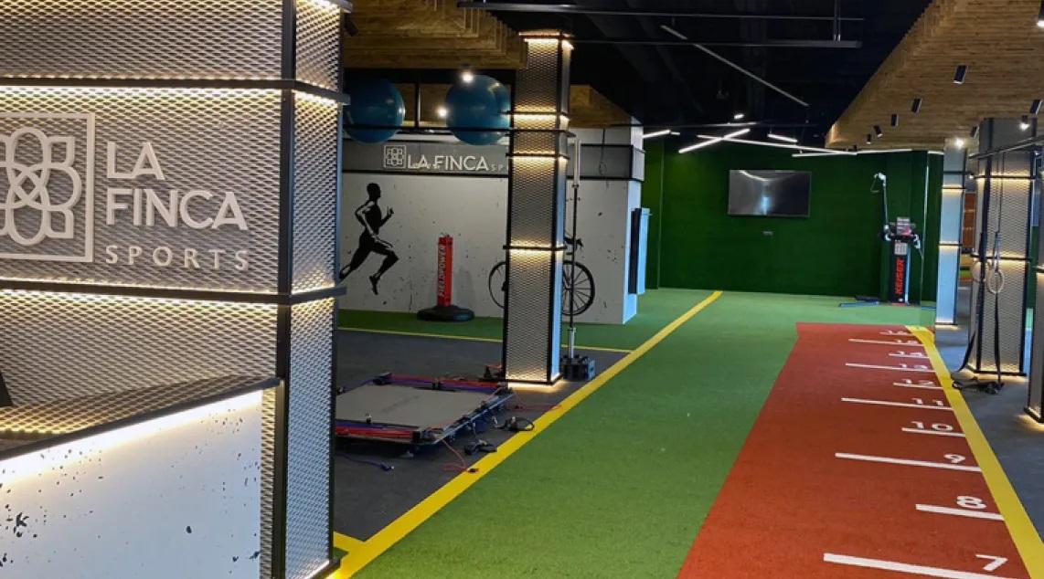 Indoor sports facility corridor with a red running track and numbered lanes, green turf, and La Finca Sports branding on the left wall