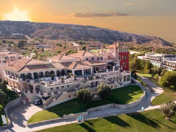 A Mediterranean-style resort on a sunlit hillside with white buildings, terraces, and palm trees at sunset.