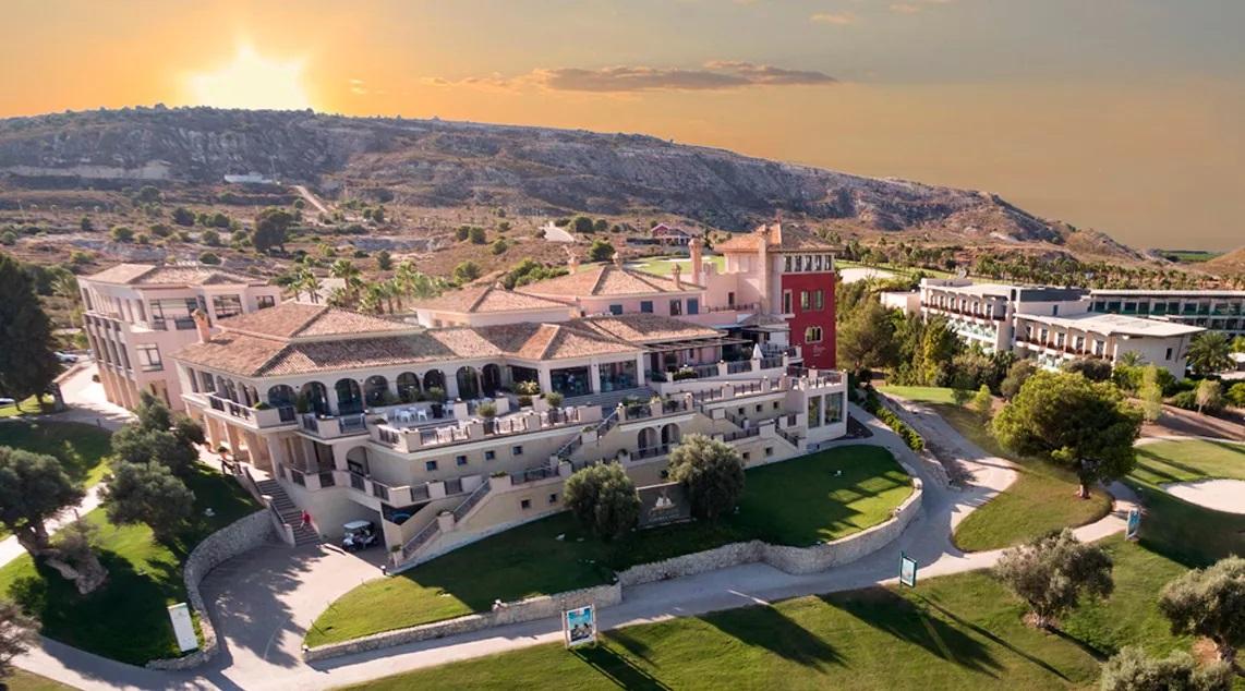 A Mediterranean-style resort on a sunlit hillside with white buildings, terraces, and palm trees at sunset.