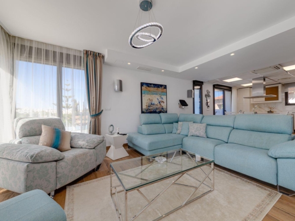 Bright coastal living room with a light blue sectional, matching armchair, glass coffee table, and sheer curtains by large windows.