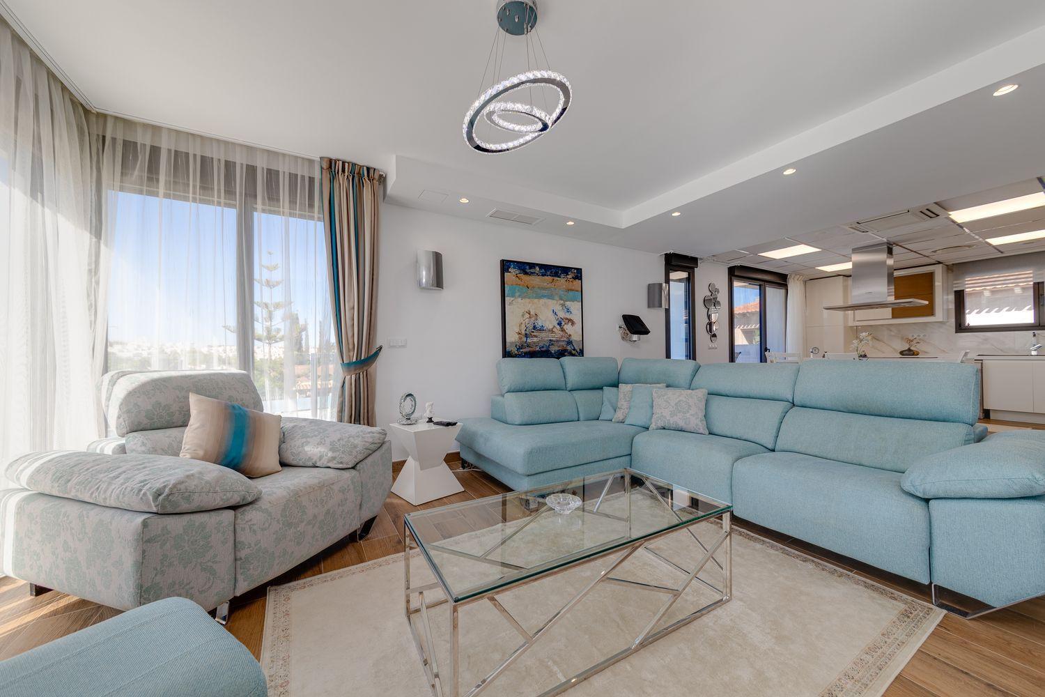 Bright coastal living room with a light blue sectional, matching armchair, glass coffee table, and sheer curtains by large windows.