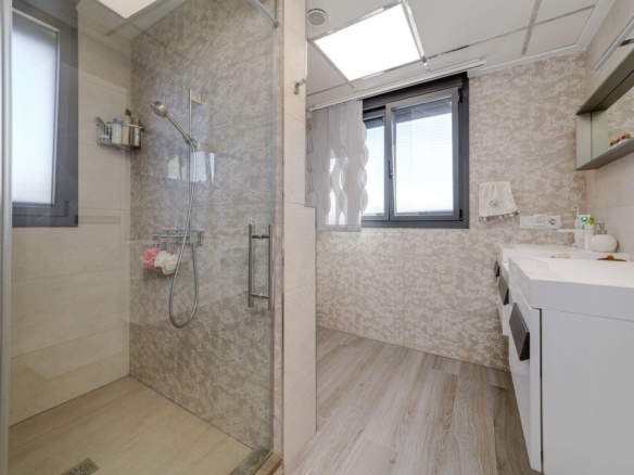 Bright modern bathroom with a glass-enclosed shower on the left, white vanity on the right, and a large window with blinds; walls have light floral-patterned tiling and wood-look flooring.