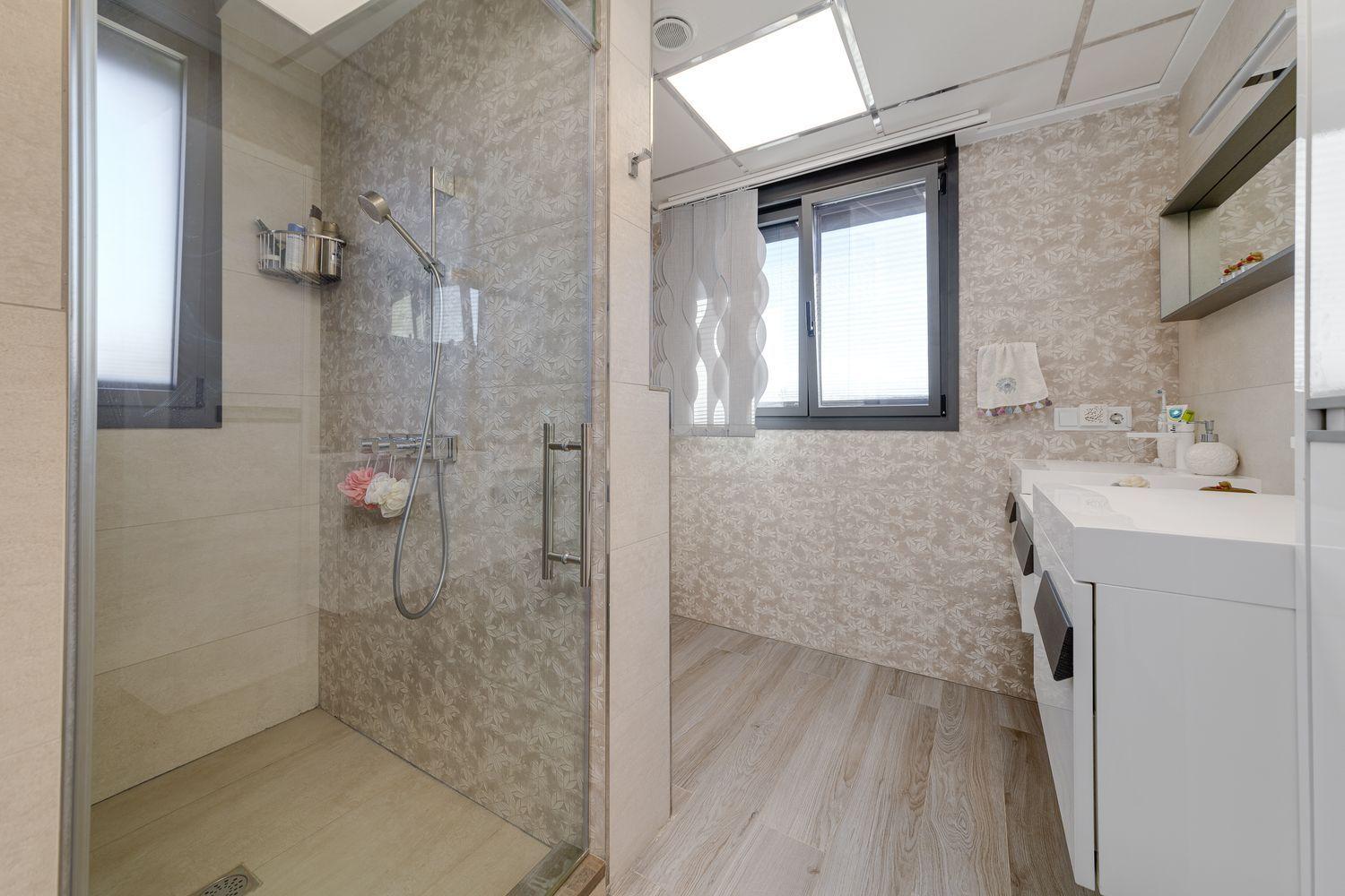 Bright modern bathroom with a glass-enclosed shower on the left, white vanity on the right, and a large window with blinds; walls have light floral-patterned tiling and wood-look flooring.