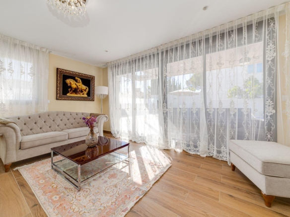 Bright, airy living room with a tufted beige sofa, glass coffee table, and lace curtains filtering natural light outside the patio doors.