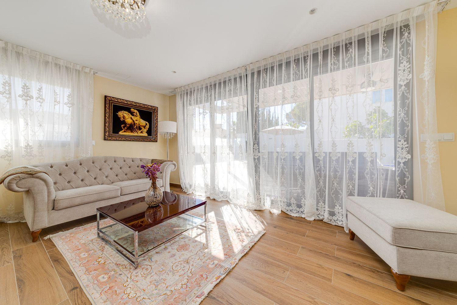 Bright, airy living room with a tufted beige sofa, glass coffee table, and lace curtains filtering natural light outside the patio doors.