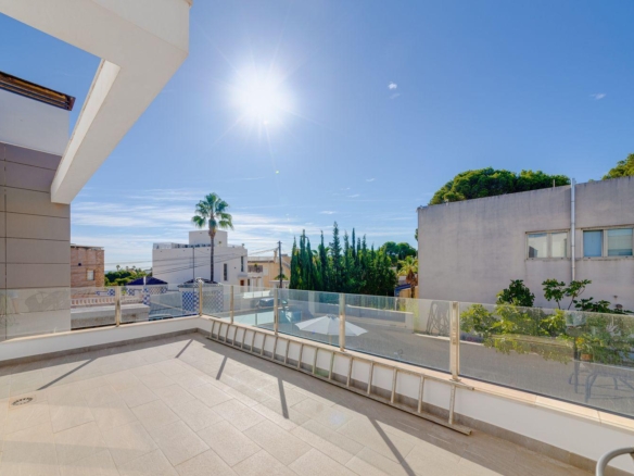 Sunny rooftop terrace with glass railing overlooking a quiet street and modern buildings under a clear blue sky