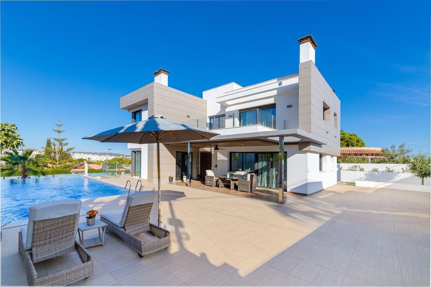 Modern white villa with beige accents, outdoor patio furniture, and a rectangular pool under a blue sky.