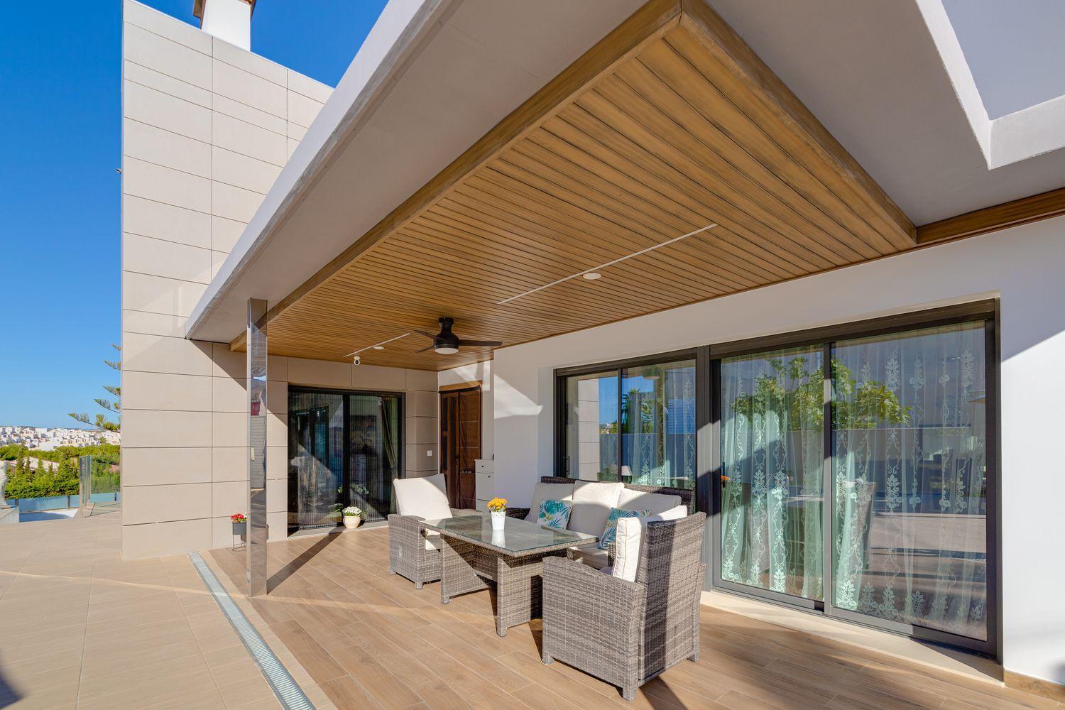 Modern outdoor patio with wicker seating around a glass-topped table under a wooden ceiling on a sunny day.