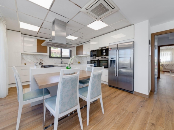 Bright modern kitchen with white cabinets, wooden accents, a central island range hood, and a wooden dining table with four upholstered chairs.