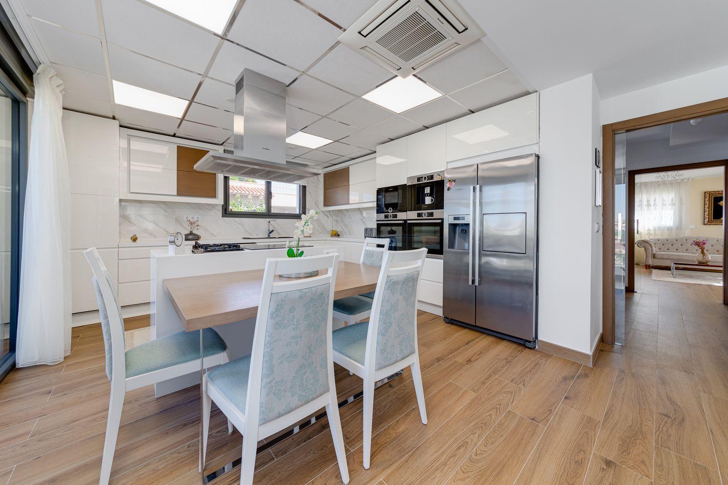 Bright modern kitchen with white cabinets, wooden accents, a central island range hood, and a wooden dining table with four upholstered chairs.