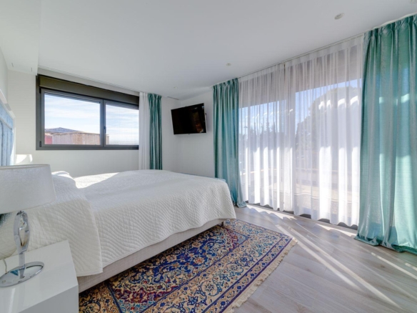 Bright hotel bedroom with white quilted bed, teal drapes, and a patterned area rug; wall-mounted TV to the right and large sliding doors letting in light.