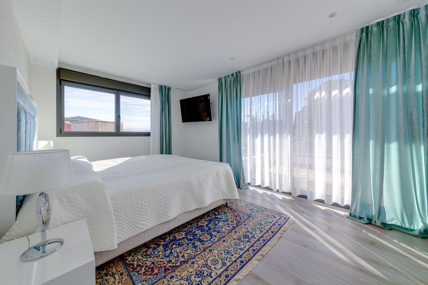 Bright hotel bedroom with white quilted bed, teal drapes, and a patterned area rug; wall-mounted TV to the right and large sliding doors letting in light.