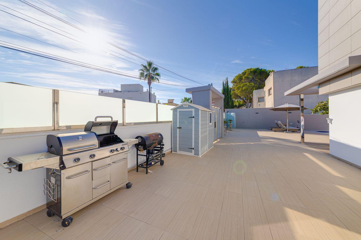 A stainless steel barbecue grill on a tiled patio with a small gray shed nearby and a bright blue sky above.