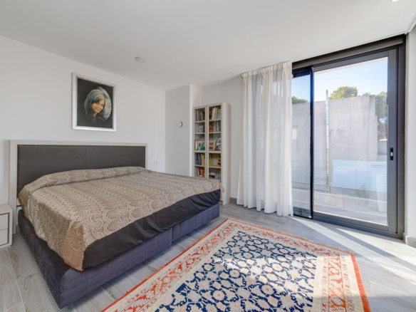Bedroom with a large upholstered bed, beige patterned duvet, and a tall headboard against a light gray wall; a white nightstand sits to the left with a salt lamp; a white bookshelf filled with books stands in the corner. A sliding glass door with sheer white curtains leads to an outdoor view, and a blue and orange patterned rug lies on the floor.