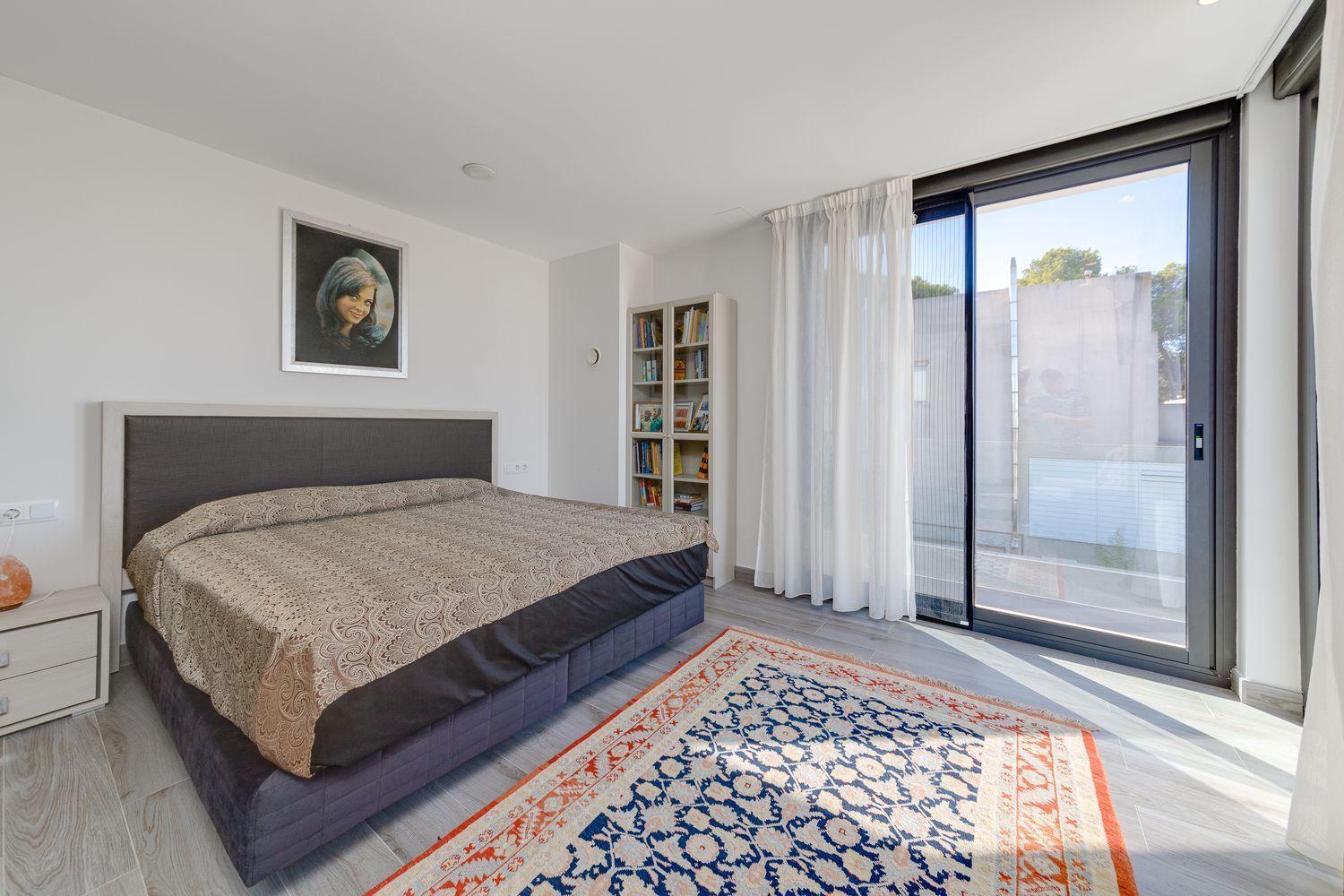 Bedroom with a large upholstered bed, beige patterned duvet, and a tall headboard against a light gray wall; a white nightstand sits to the left with a salt lamp; a white bookshelf filled with books stands in the corner. A sliding glass door with sheer white curtains leads to an outdoor view, and a blue and orange patterned rug lies on the floor.