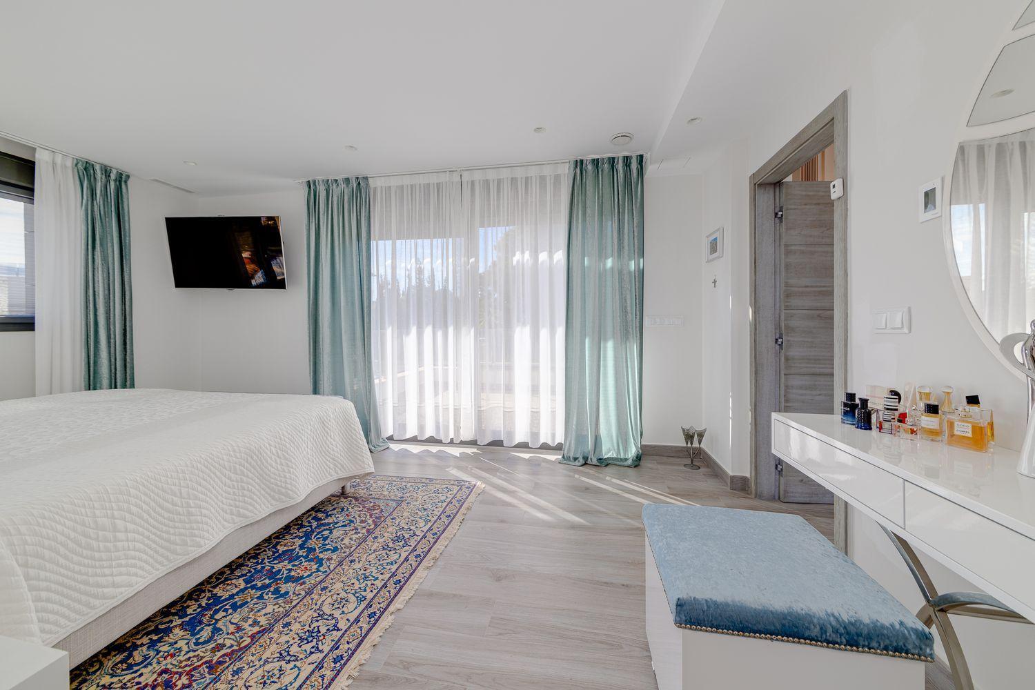 Bright hotel-style bedroom with a white quilted bed, teal drapes, and sheer curtains by large windows