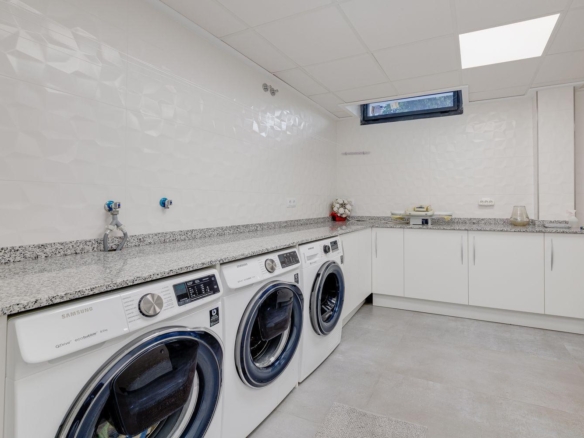 Bright laundry room with three white front-loading washing machines under a speckled granite counter and white wall tiles with a small high window nearby.