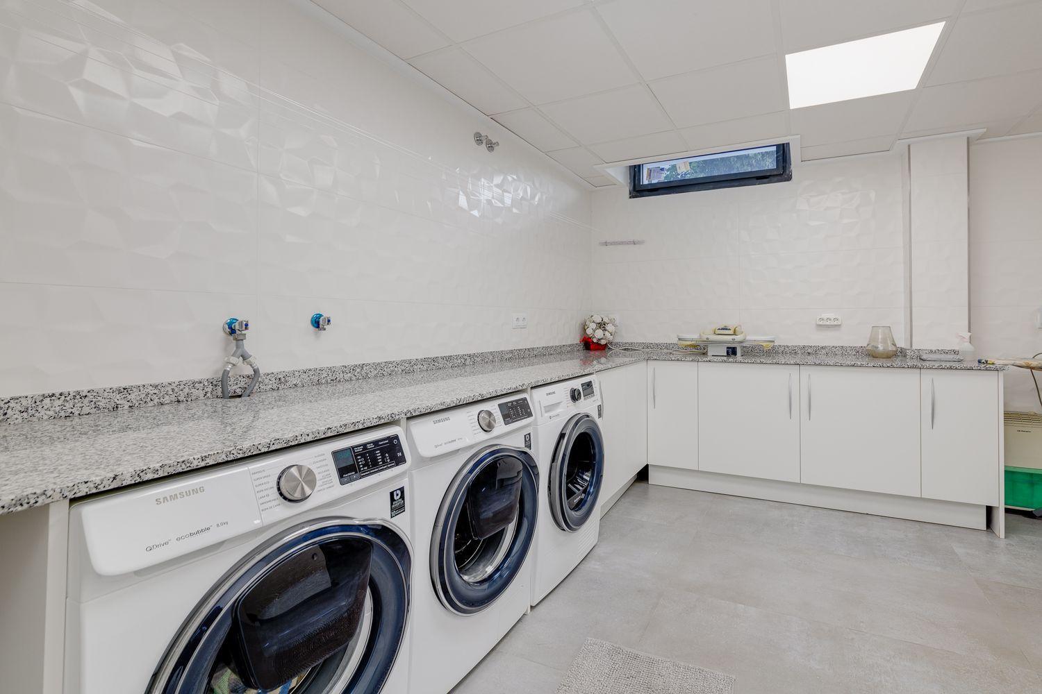 Bright laundry room with three white front-loading washing machines under a speckled granite counter and white wall tiles with a small high window nearby.