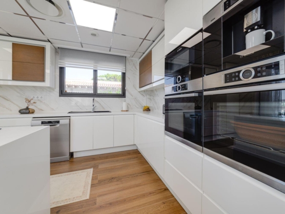Modern white kitchen with glossy cabinets, built-in Bosch ovens, and a marble backsplash under bright ceiling panels, plus a window over the sink.
