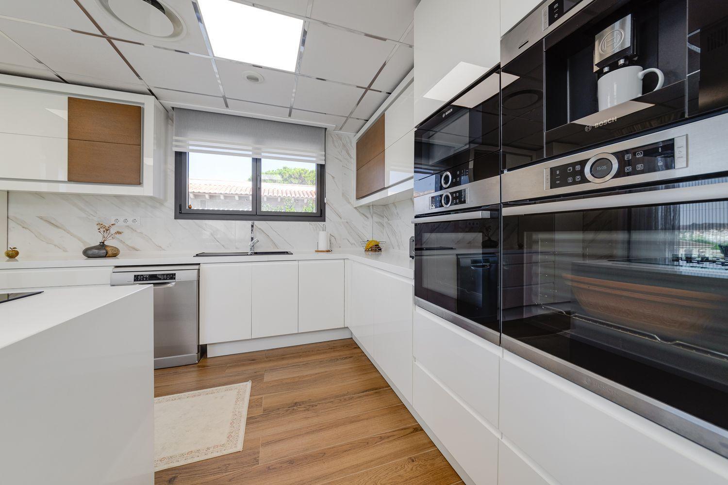 Modern white kitchen with glossy cabinets, built-in Bosch ovens, and a marble backsplash under bright ceiling panels, plus a window over the sink.