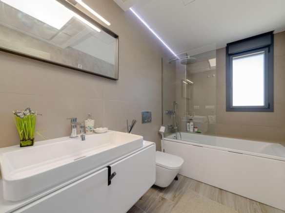 Contemporary bathroom with a white vanity and rectangular sink, mirror, and chrome fixtures, beige tiled walls, a glass shower screen, and a built-in bathtub with a rainfall showerhead.