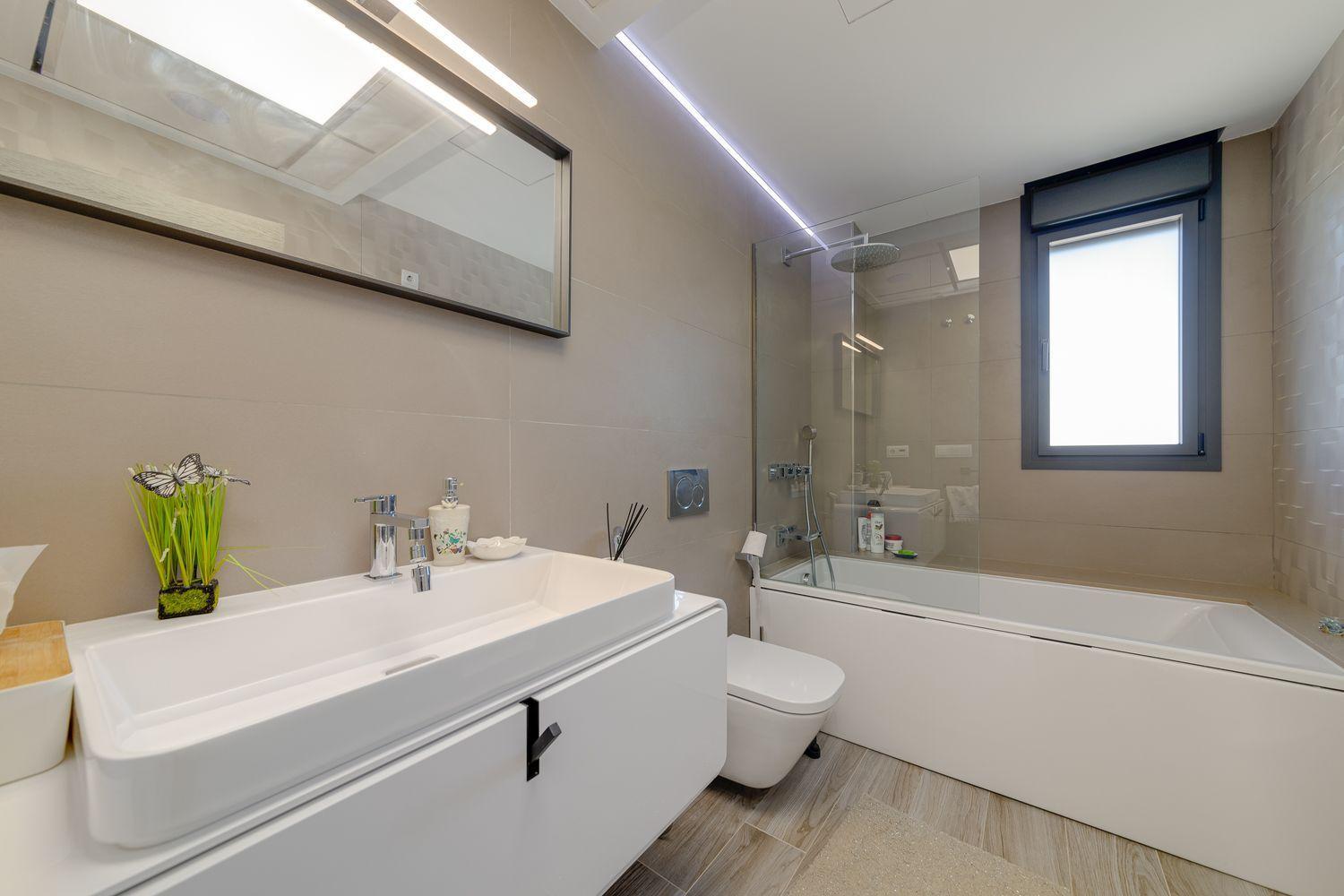 Contemporary bathroom with a white vanity and rectangular sink, mirror, and chrome fixtures, beige tiled walls, a glass shower screen, and a built-in bathtub with a rainfall showerhead.
