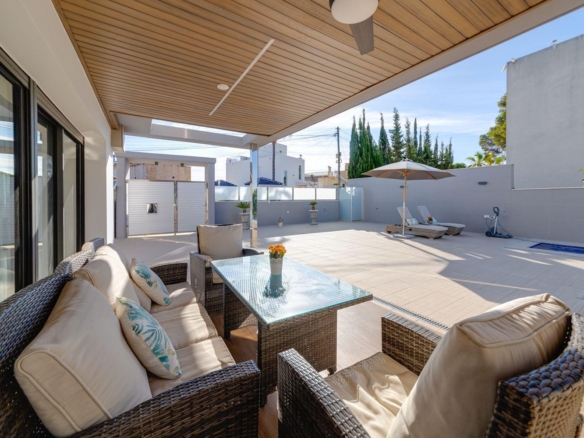 Cozy outdoor seating area on a covered patio with beige cushions, wicker furniture, and a glass-topped table, beside a sunny pool area.