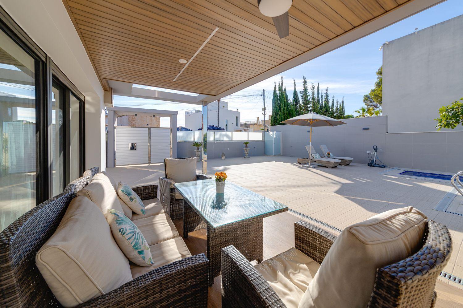 Cozy outdoor seating area on a covered patio with beige cushions, wicker furniture, and a glass-topped table, beside a sunny pool area.