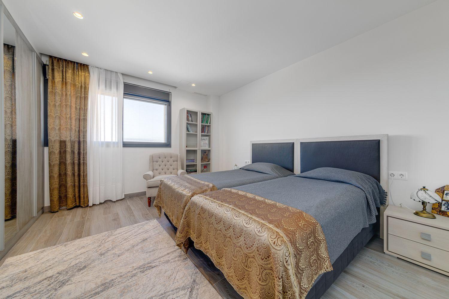 Bedroom with two twin beds side by side, blue upholstered headboards, and gold-patterned bed runners; window with sheer white and gold curtains nearby.