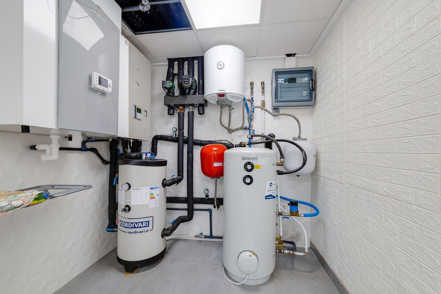 Utility room with multiple water heaters and an expansion tank, exposed pipes and a control panel on the wall