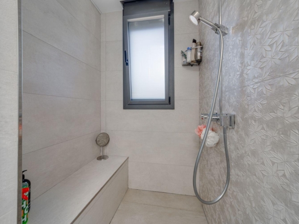 Modern shower stall with light gray tiles, a built-in bench, frosted window, and a handheld chrome showerhead with a hanging hose and caddy of toiletries.