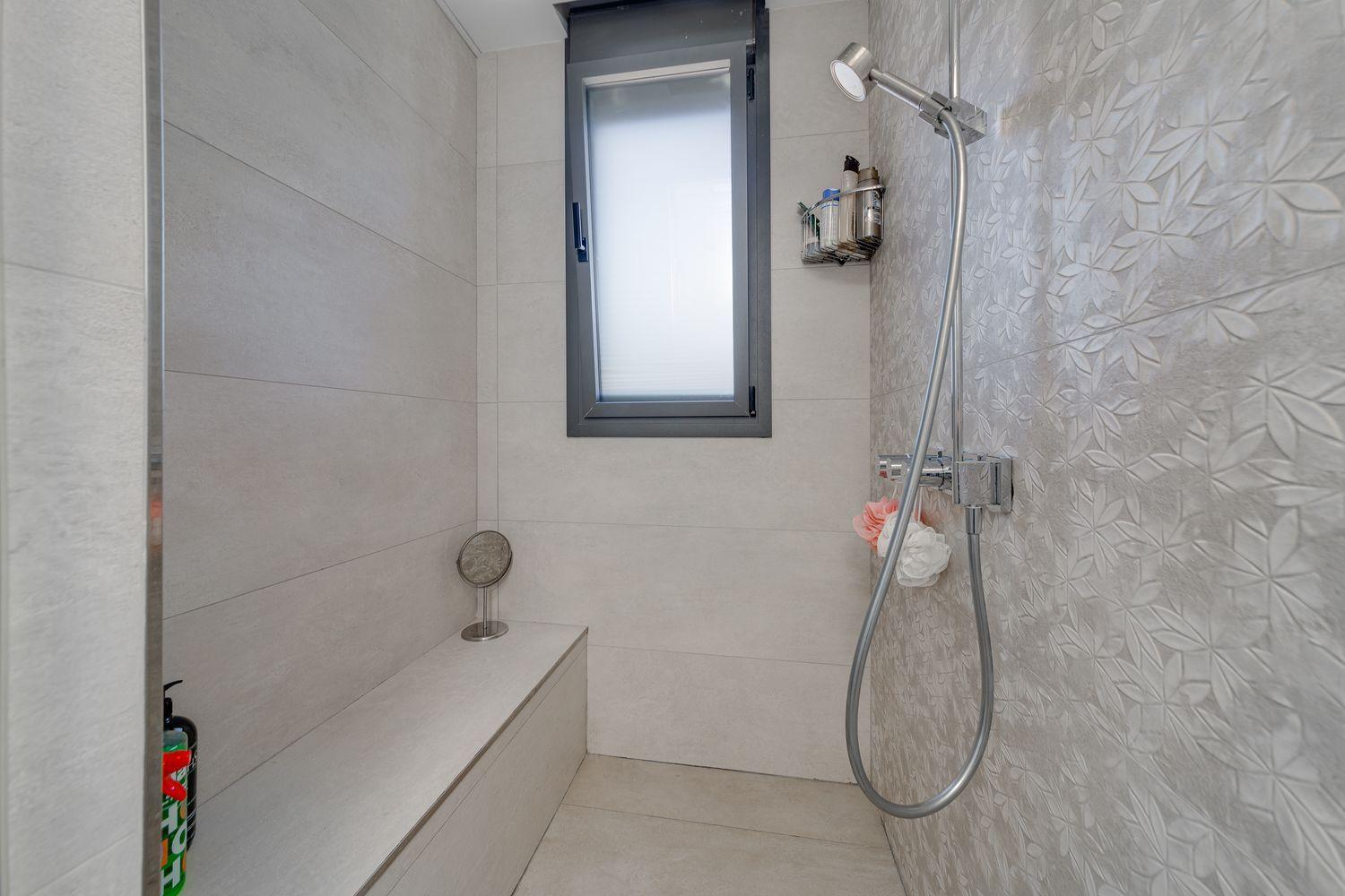Modern shower stall with light gray tiles, a built-in bench, frosted window, and a handheld chrome showerhead with a hanging hose and caddy of toiletries.