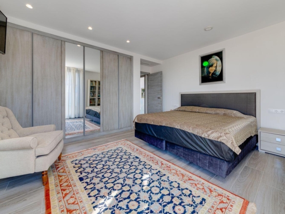 Modern bedroom with a king bed, tufted headboard, mirrored wardrobe doors, and a beige armchair.