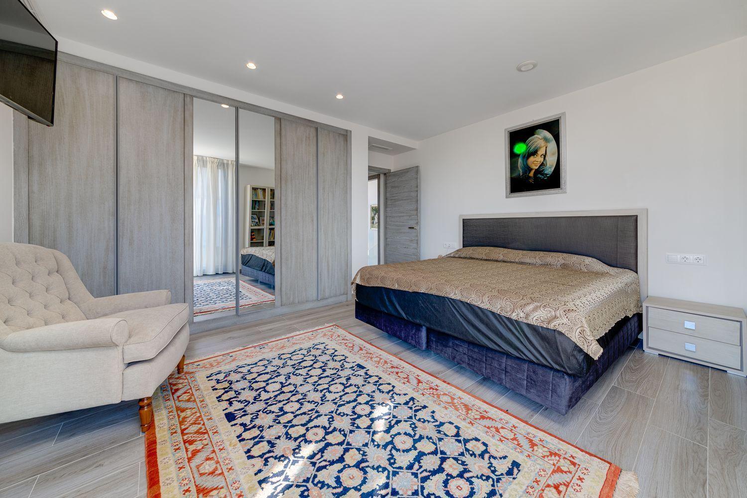 Modern bedroom with a king bed, tufted headboard, mirrored wardrobe doors, and a beige armchair.